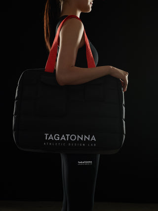 Woman  wearing a black TAGATONNA Pilates tote bag with reflective branding on a black background