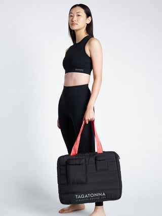 Woman  wearing a black TAGATONNA Pilates tote bag with reflective branding on a white background