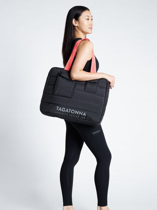 Woman  wearing a black TAGATONNA Pilates tote bag with reflective branding on a white background
