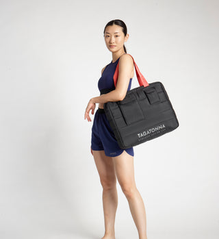 Woman  wearing a black TAGATONNA Pilates puffer bag with reflective branding on a white background